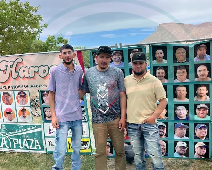 Three young men stand together in front of the Faces of the Harvest wall, a chain-link fence hung with portraits of farmworkers, with a La Plaza banner and Mount Garfield behind