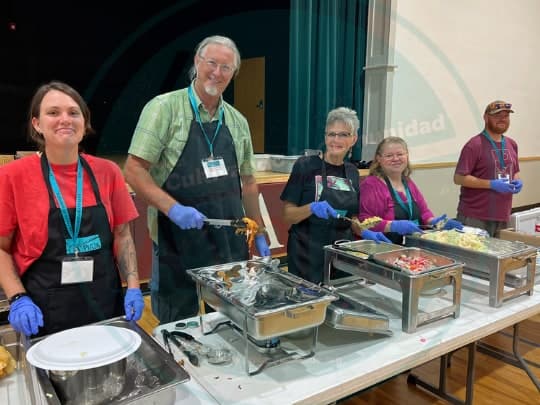 Volunteers in aprons and blue gloves smile behind trays of food at La Plaza's Farewell Dinner